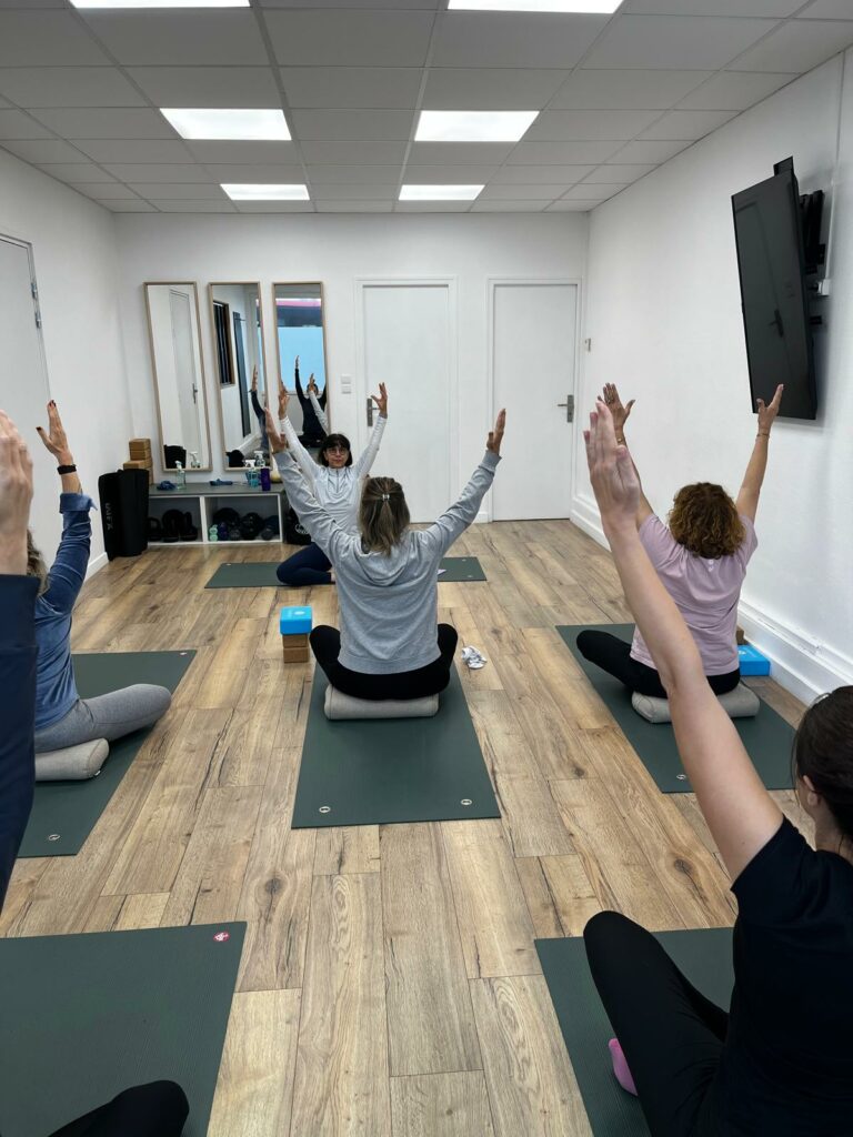 Cours collectif de yoga : élèves assis sur des coussins, bras levés, face à la professeure dans un studio blanc au parquet
