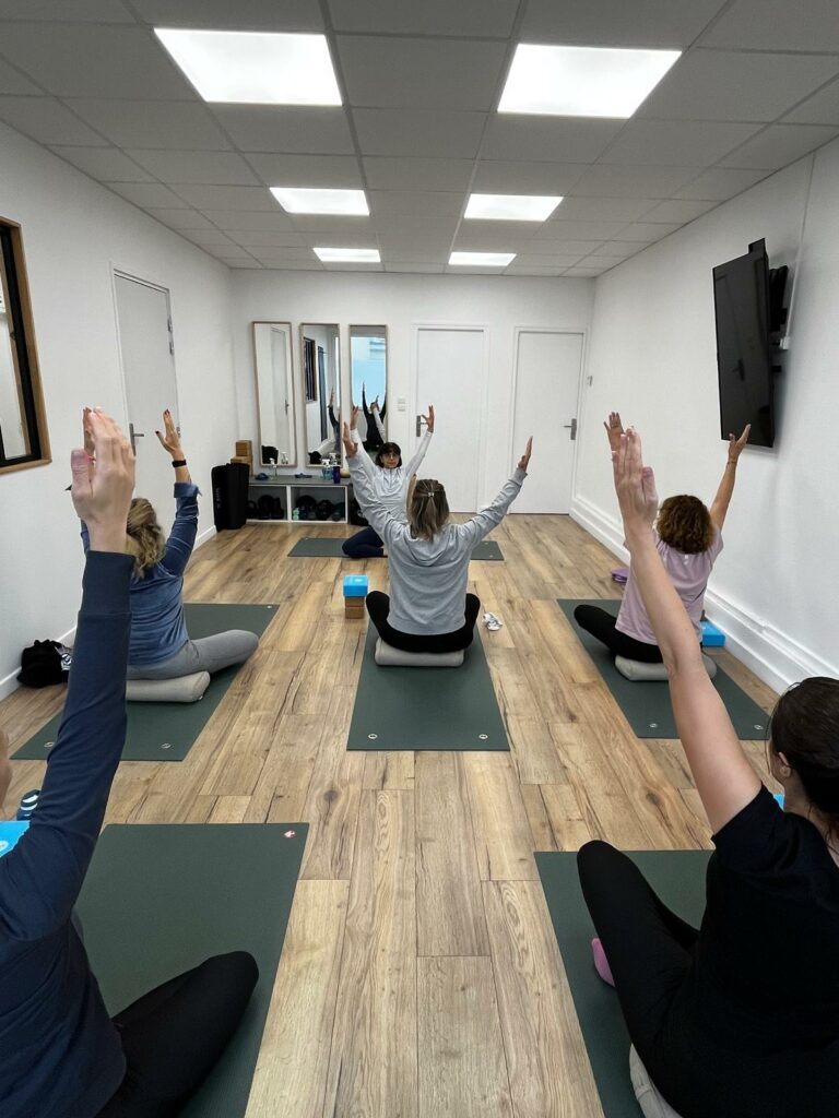 Élèves de yoga assis, bras levés pendant un cours collectif, la professeure au fond du studio blanc