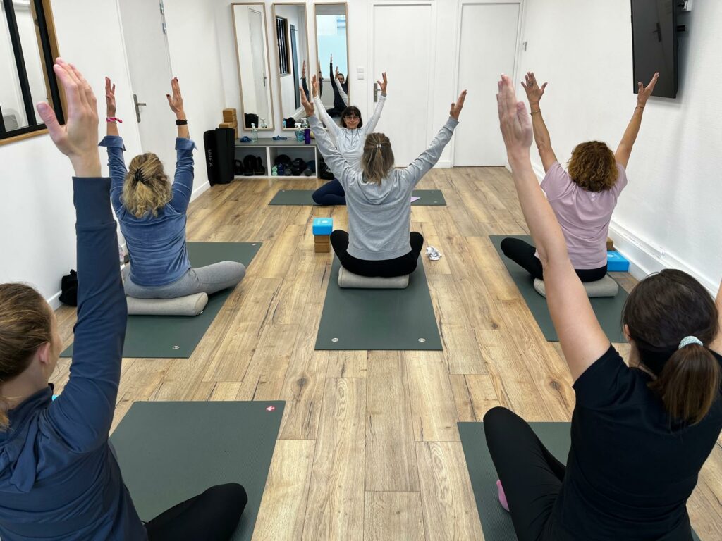 Cours de yoga collectif, bras levés vers le ciel, élèves assis sur des tapis verts dans un studio blanc au parquet