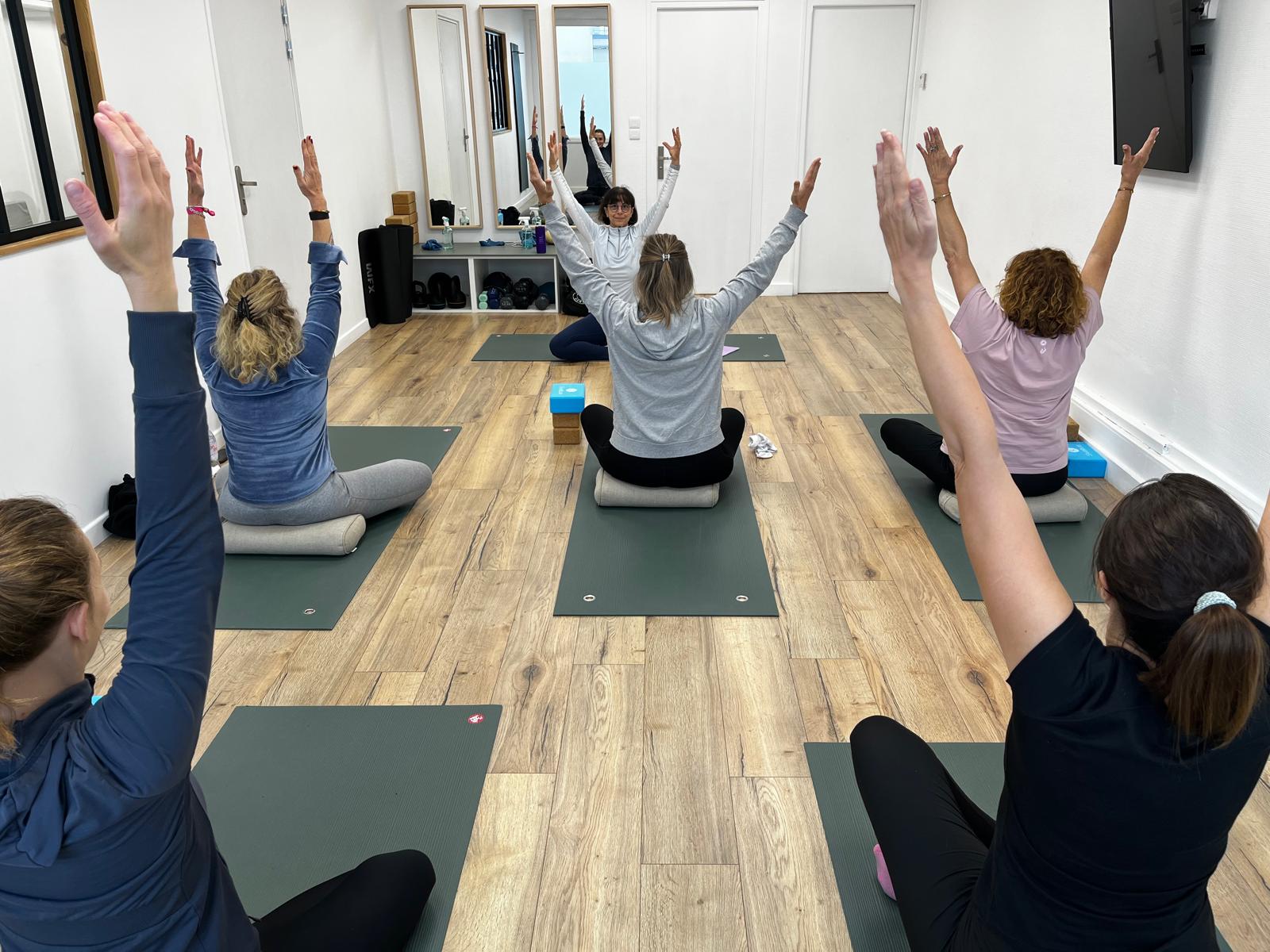 Cours de yoga collectif, bras levés vers le ciel, élèves assis sur des tapis verts dans un studio blanc au parquet